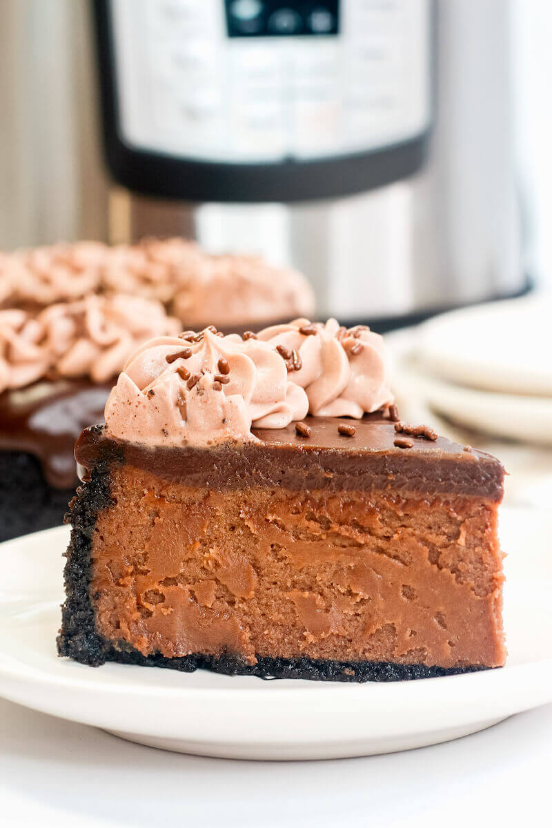 Side view of a slice of nutella cheesecake in front of the whole cheesecake and an instant pot.