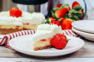 A white plate with a slice of strawberry cheesecake with whipped cream topping, next to a ripe red strawberry.