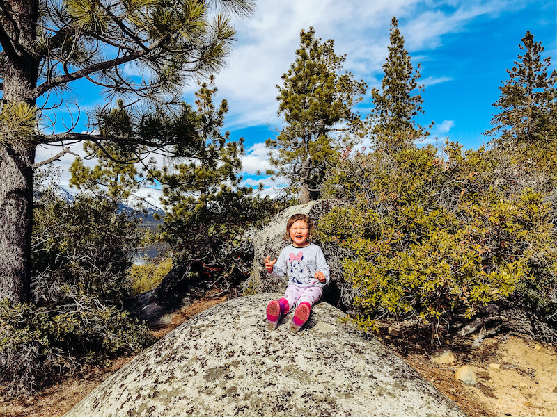 A little girl sitting on a rock surrounded by trees and smiling.