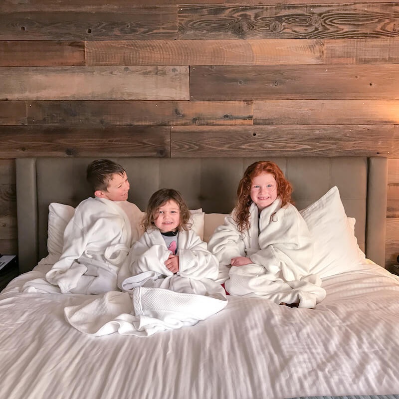 Three kids in white hotel robes sitting on a king sized bed with white bedding.