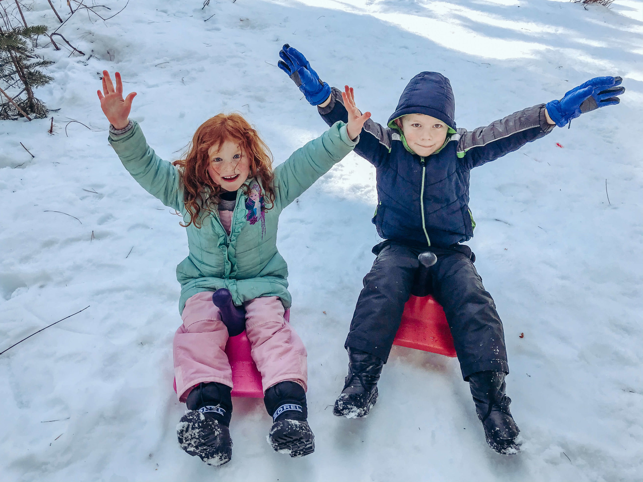 Two kids sitting on sleds with their arms up, surrounded by snow.