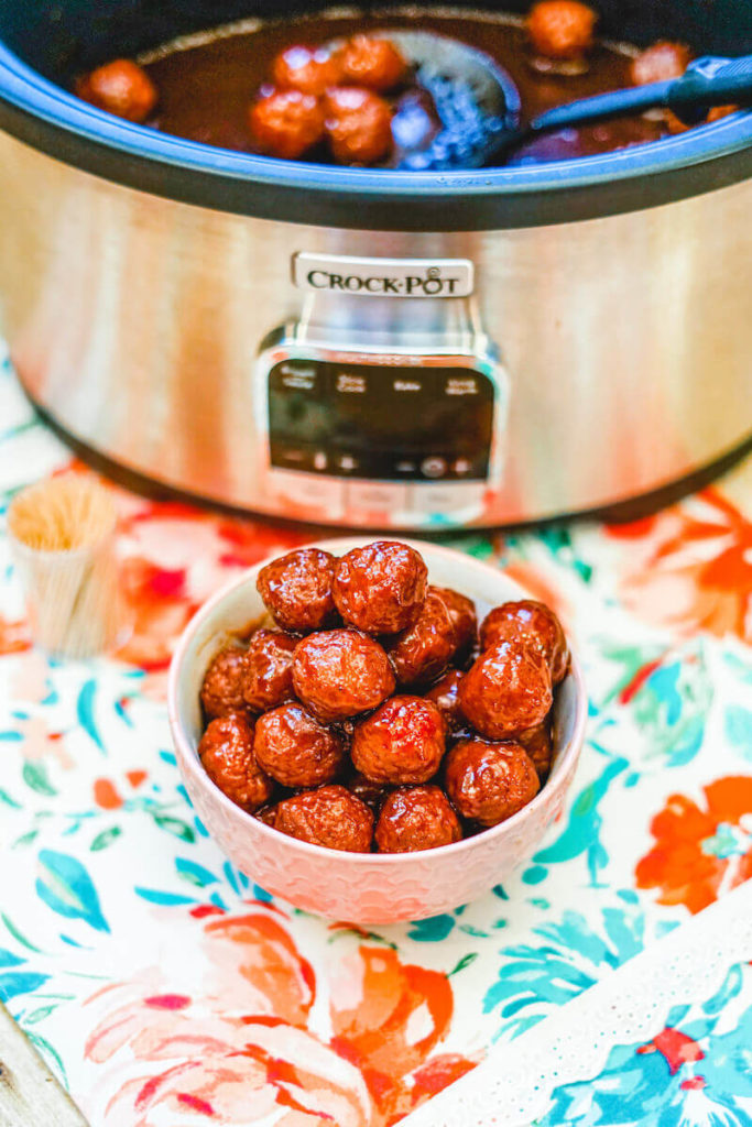 A small white bowl filled with grape jelly meatballs in front of a crock pot.