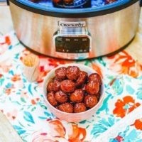 A crockpot on a floral placemat with a bowl of grape jelly meatballs in a white bowl.