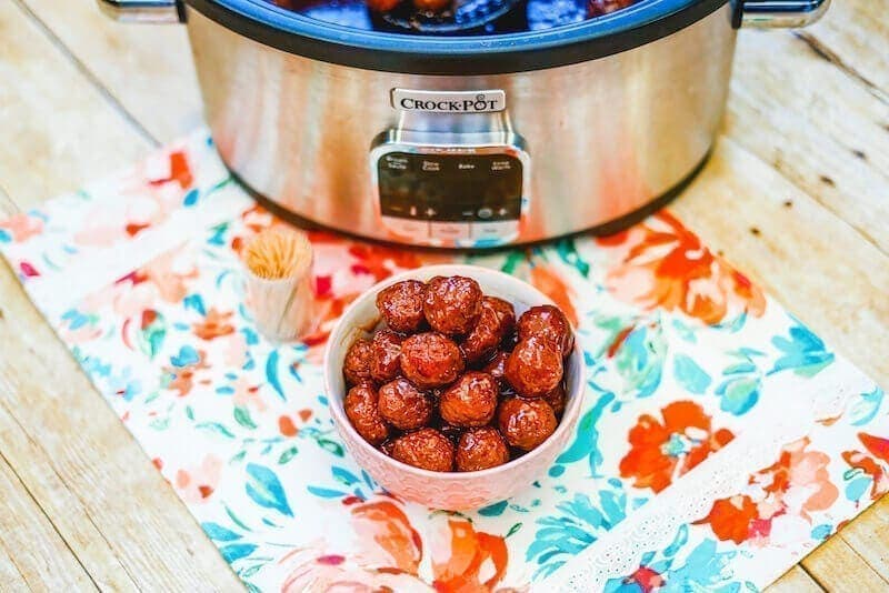 A crockpot on a floral placemat with a bowl of grape jelly meatballs in a white bowl.