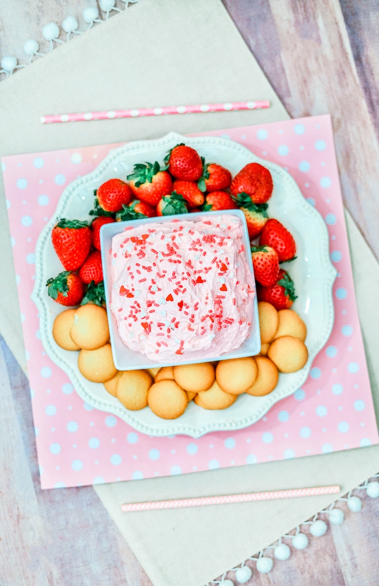 A pink and white polka dot placemat filled with a platter of strawberry cake batter dip, strawberries, and cookies.