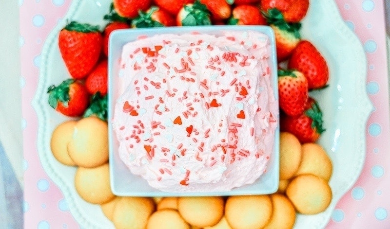 A white plate filled with strawberries, vanilla cookies, and a square bowl of strawberry cake batter dip in the center.