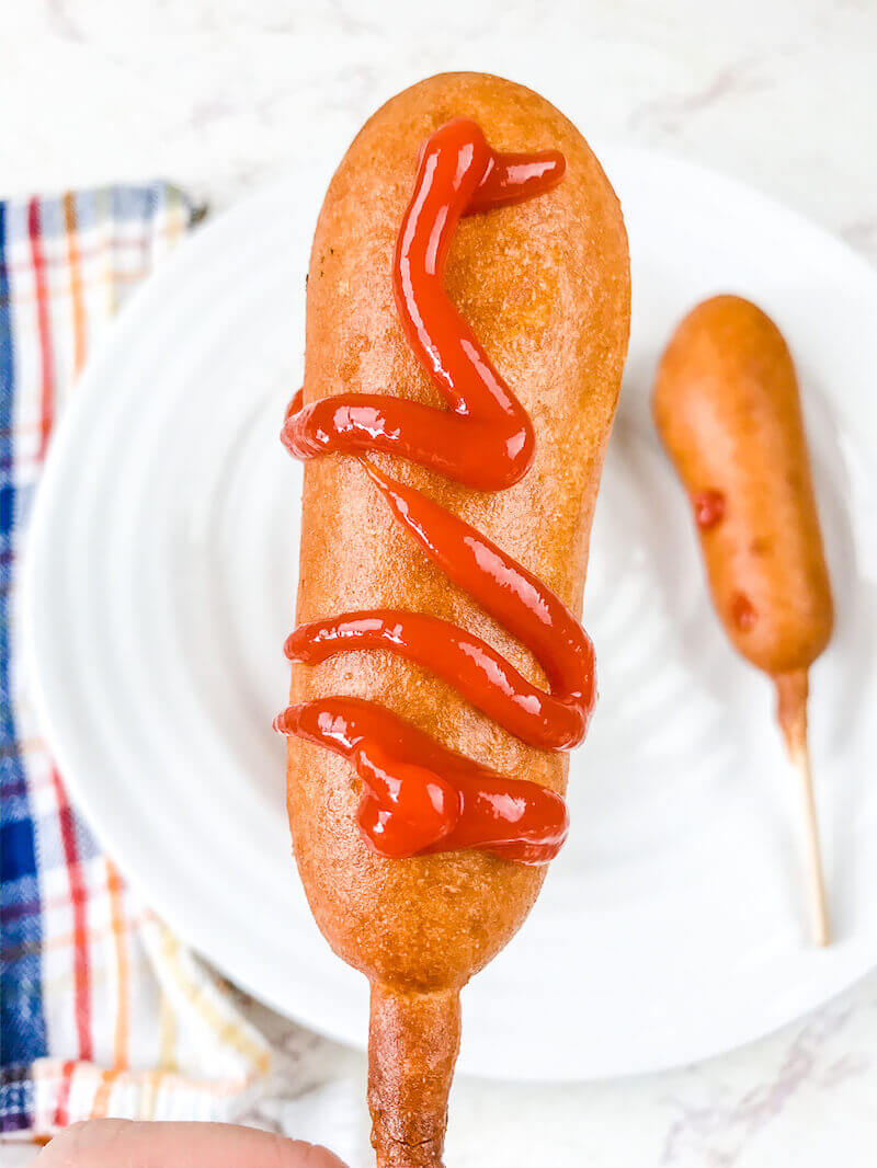 A hand holding up a corn dog with ketchup on it.
