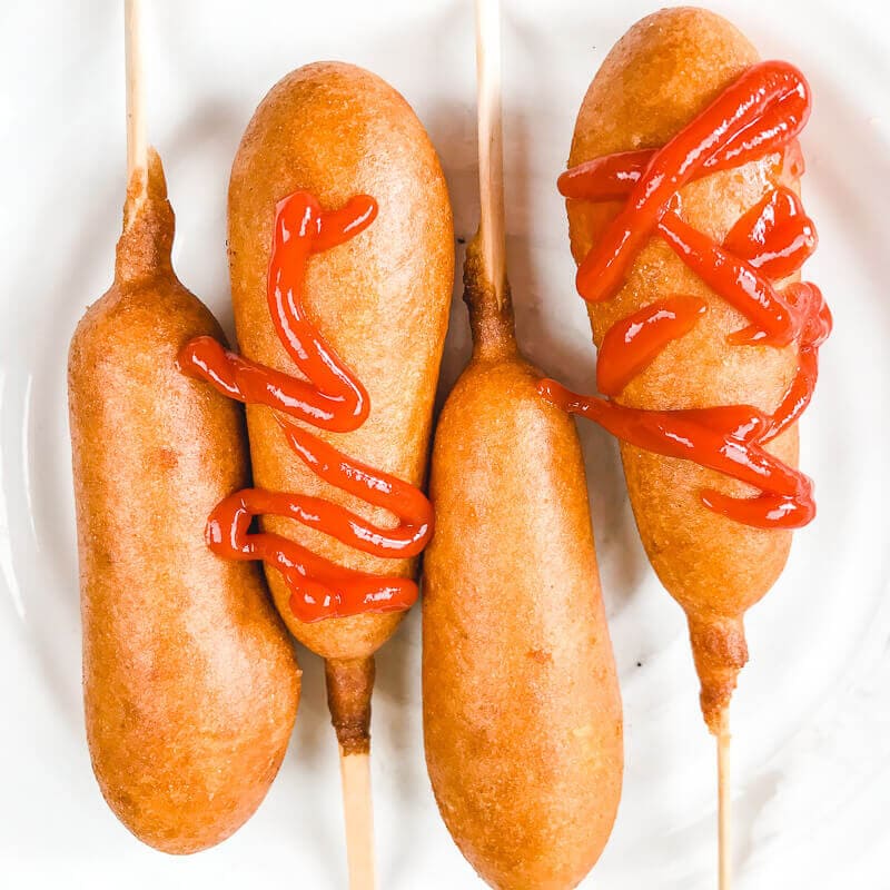 A white plate with four corn dogs, two are drizzled with ketchup.