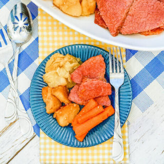 A blue checkered place mat topped with a plate of corned beef with potatoes and carrot.