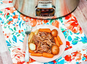 A plate of roast beef with carrots and potatoes in front of a crockpot.