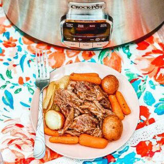 A plate of roast beef with carrots and potatoes in front of a crockpot.