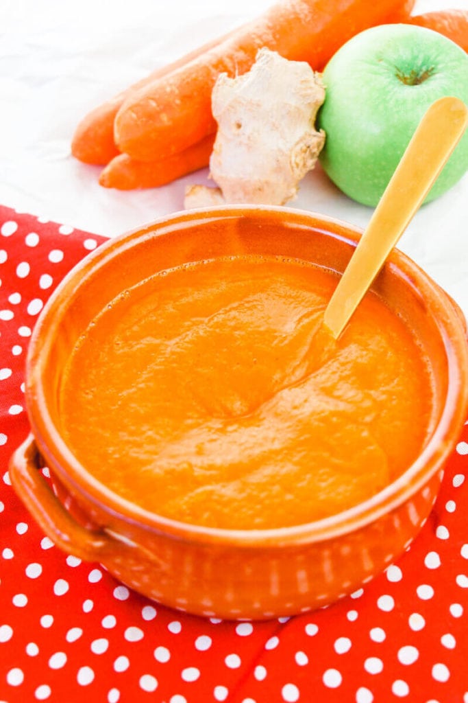 A bowl of carrot soup with carrots, apples, and ginger in the background.