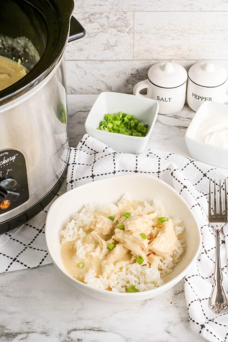 A crockpot with the lid off next to a white bowl filled with chicken and gravy over rice and a small bowl of diced green onions.
