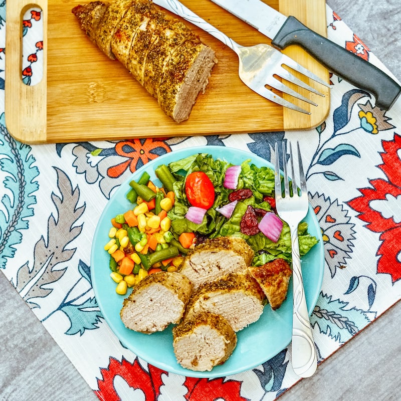 A blue plate loaded with sliced pork tenderloin and cooked vegetables next to a cutting board with pork tenderloin on a steak knife.