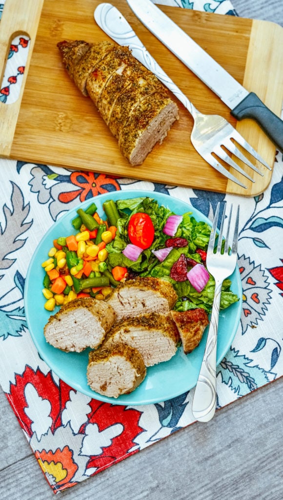 A floral tablecloth with a plate of pork tenderloin with mixed veggies next to a cutting board with a pork tenderloin cut in half.