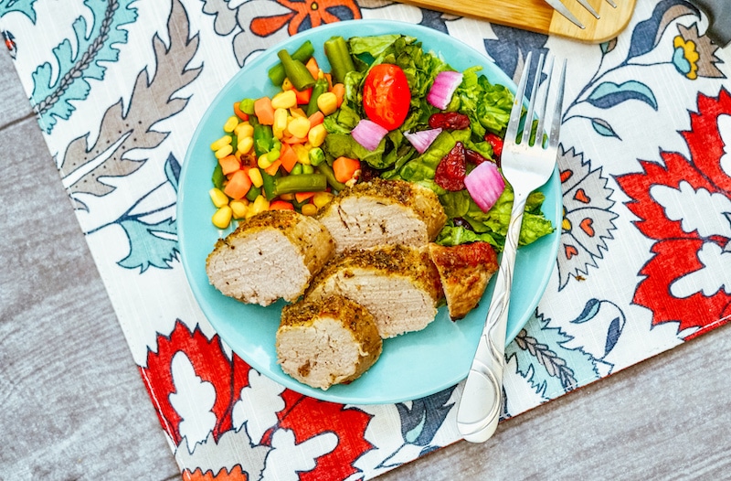 A blue plate of slices of pork tenderloin next to a green salad and mixed veggies.