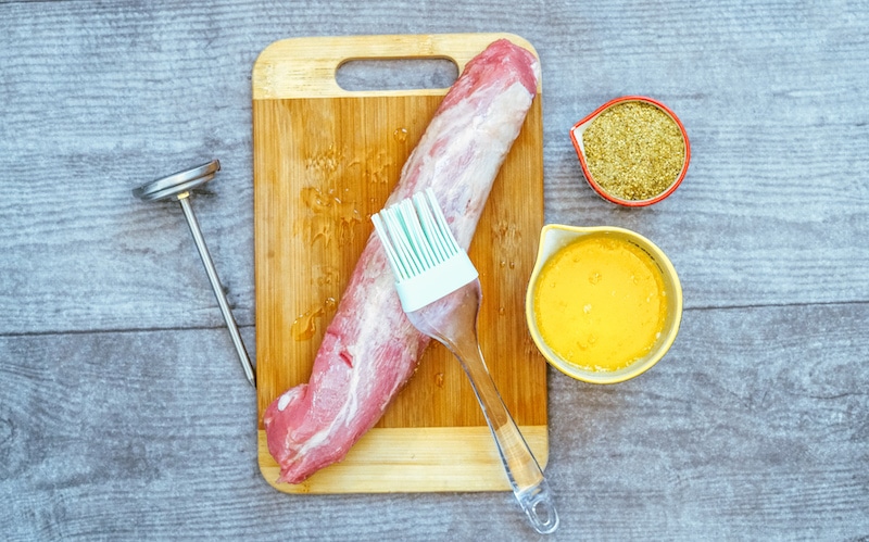 A raw pork tenderloin on a butting board being brushed with melted butter.
