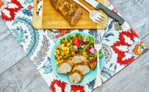 A floral placemat with a cutting board topped with pork tenderloin and a light blue plate of pork and vegetables.