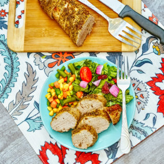 A floral placemat with a cutting board topped with pork tenderloin and a light blue plate of pork and vegetables.