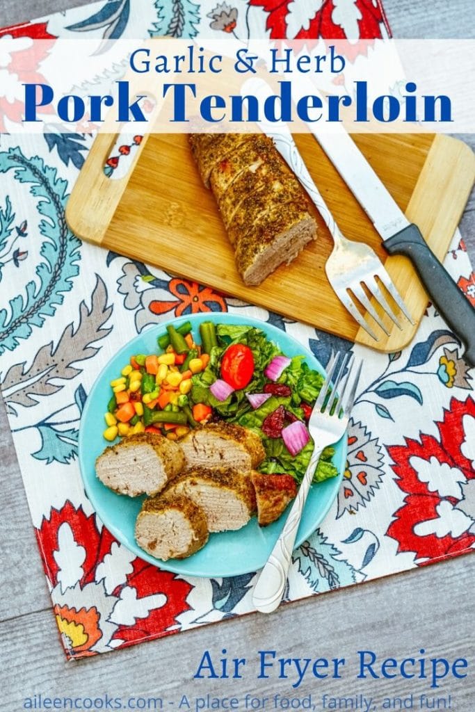 A plate of pork tenderloin over a floral placemat with the words "garlic & herb pork tenderloin" in blue lettering.