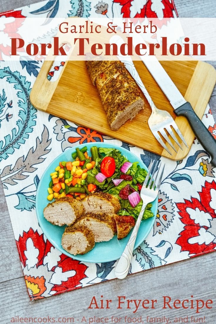 A plate of pork tenderloin over a floral placemat with the words "garlic & herb pork tenderloin" in red lettering.