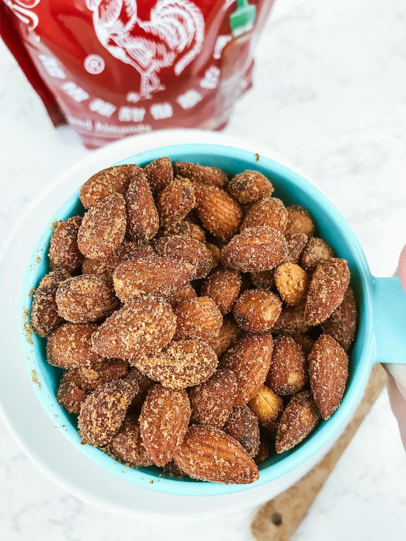 A blue measuring cup holding up a heaping cup full of sriracha spiced almonds.