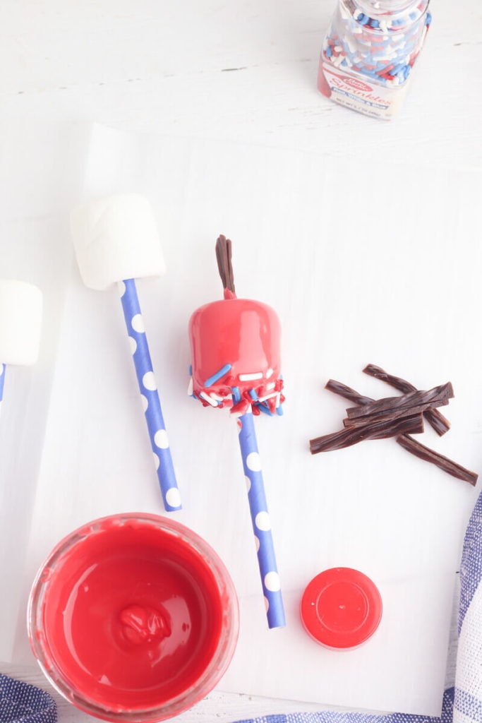 A plain marshmallow on a straw next to a red dipped marshmallow with black licorice sticking out the top and red, white, and blue sprinkles on the bottom.