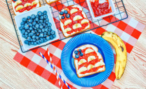 A piece of toast decoarated to look like an American flag next to a banana, bowl of blueberries, and bowl of strawberry jam.