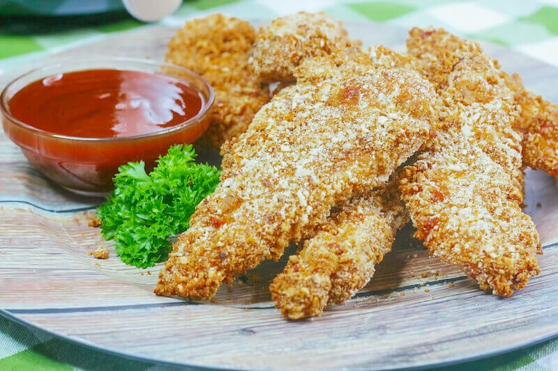 Side view of a plate of air fried chicken strips next to a small dish of ketchup.
