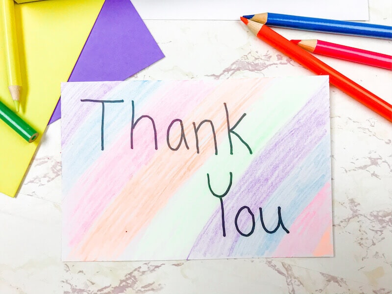 A rainbow colored card with the words Thank You next to a pile of colored pencils.