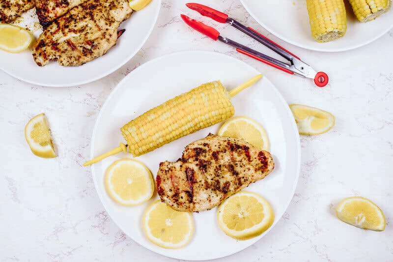 A white plate of grilled chicken, sliced lemons, and corn on the cob.