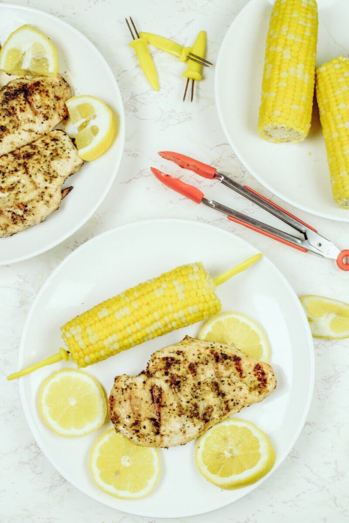 A plate of grilled chicken next to a plate of corn on the cob.