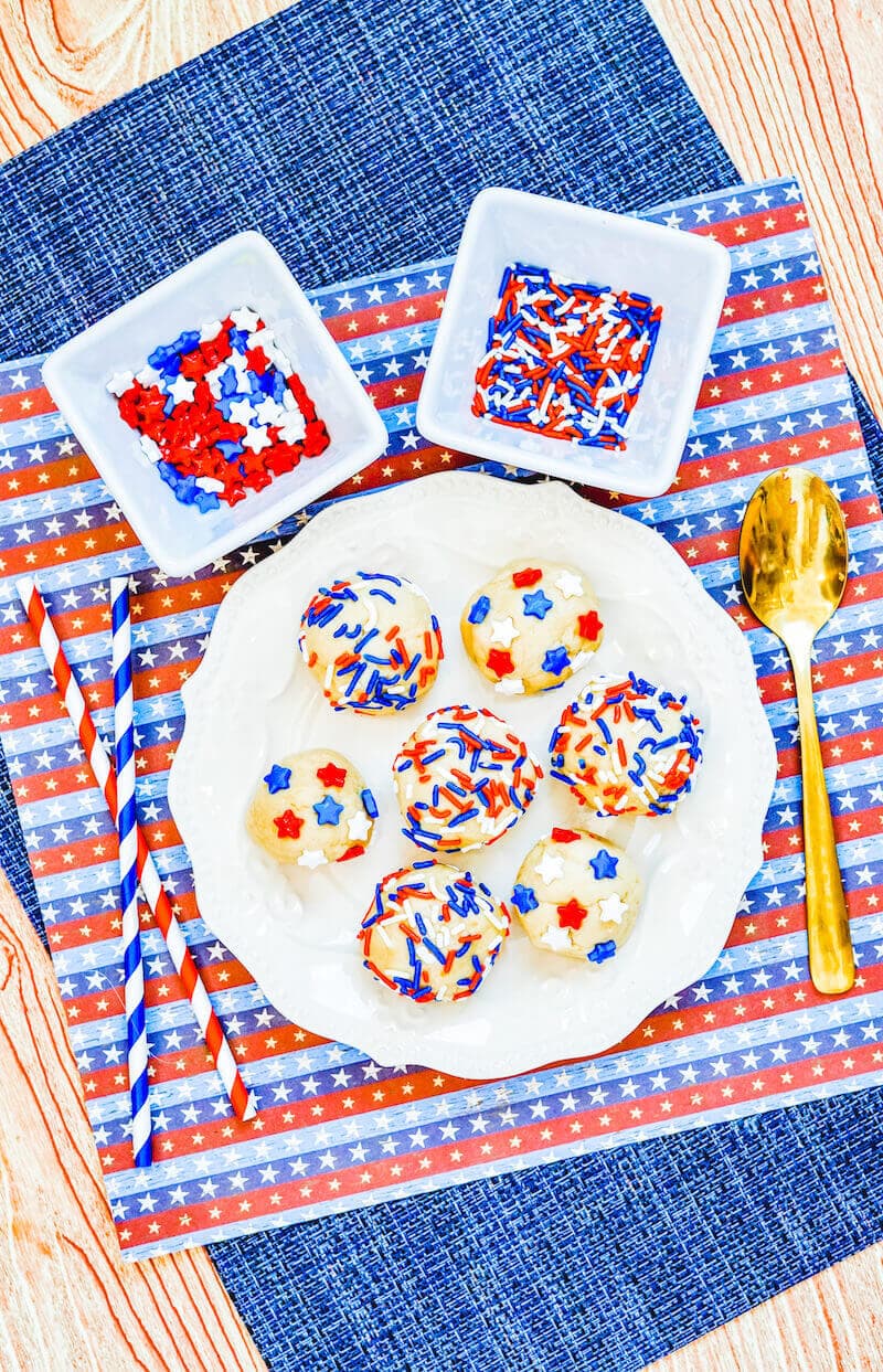 Overhead shot of edible sugar cookie dough rolled into balls on a white plate, next to a bold spoon and bowls of sprinkles.