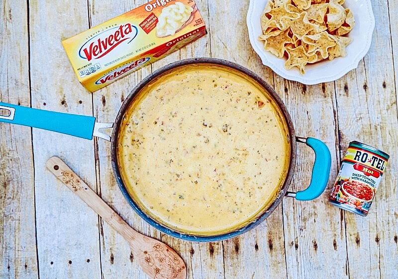 A blue sauce pan filled with fully cooked Rotel cheese dip next to a box of Velveeta and a plate of tortilla chips.