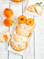 A white serving tray filled with peach hand pies and next to a peach cut in half.