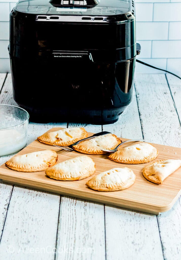 A wooden cutting board filled with hand pies, in front of a black air fryer.