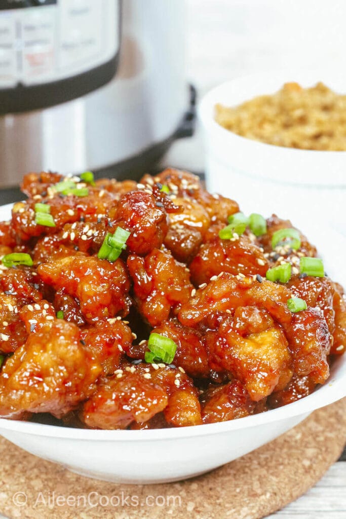A bowl of general tso's chicken in front of a small bowl of brown rice and an instant pot.