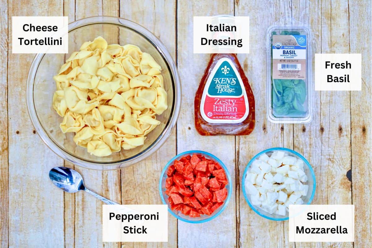 A large glass bowl of tortellini next to a bottle of salad dressing and other ingredients for pasta salad.
