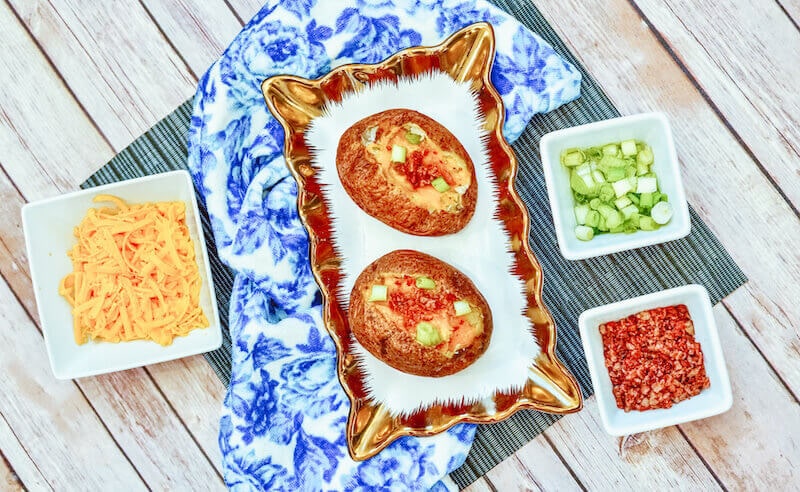 A gold and white serving dish filled with loaded baked potatoes on top of a blue and white dish towel.