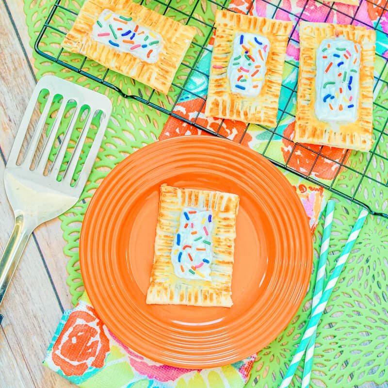 An orange plate with a pop tart on it next to a cooling rack of pop tarts.