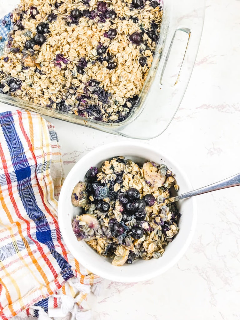 A white bowl of blueberry baked oatmeal below a baking dish of baked oatmeal.