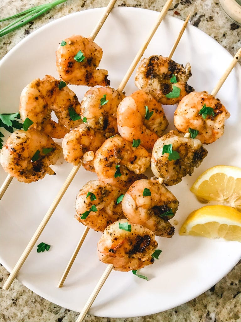 A white plate of grilled shrimp on skewers and topped with chopped parsley.