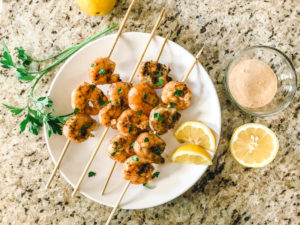 A white plate of grilled shrimp next to a sprig of Italian parsley, sliced lemons, and a small clear jar of remoulade sauce.