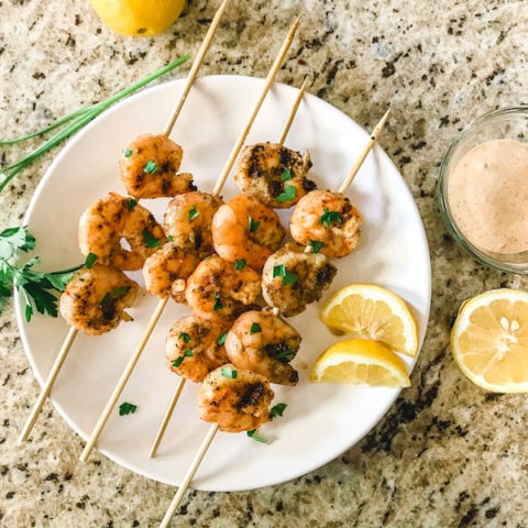 A white plate of grilled shrimp next to a sprig of Italian parsley, sliced lemons, and a small clear jar of remoulade sauce.