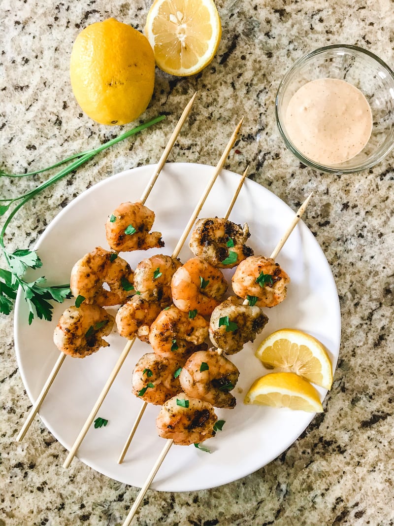 Glass bowl of Remoulade sauce above a white plate of skewered grilled shrimp.