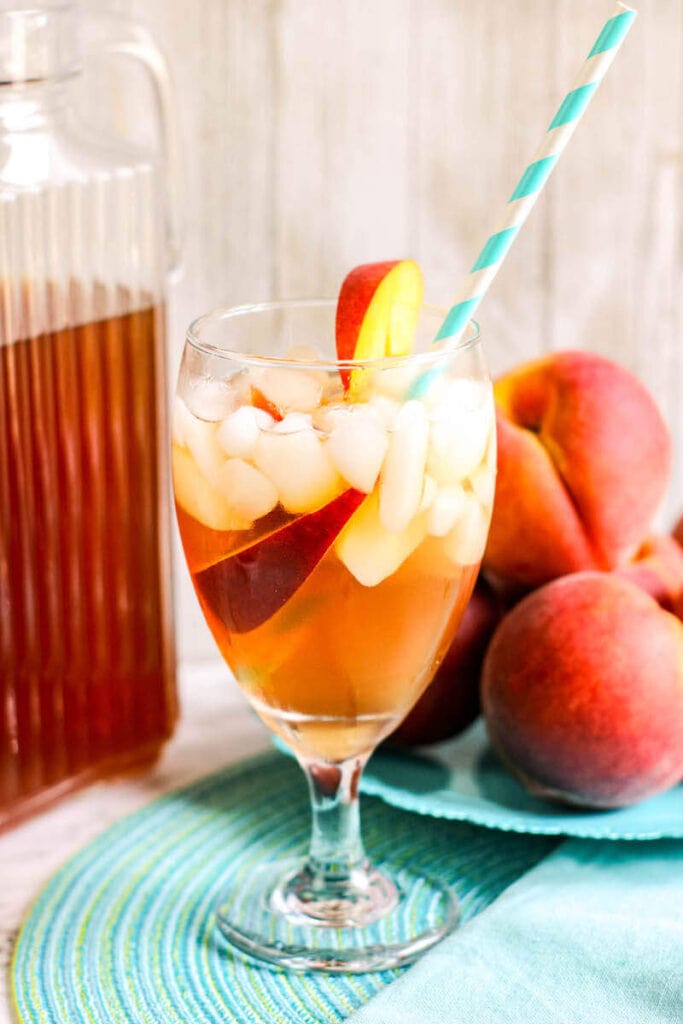 A glass of iced tea in front of a bowl of peaches and a pitcher of iced ta.