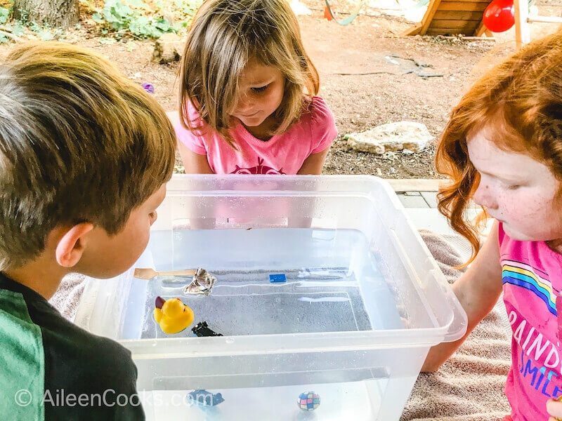 Three kids looking into a clear bin of water and various items.