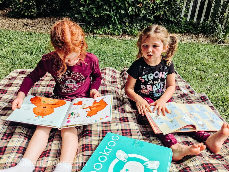 Two girls looking at picture books outside on a picnic blanket.