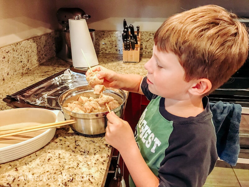 A boy adding chunks of chicken to a wooden skewer. 