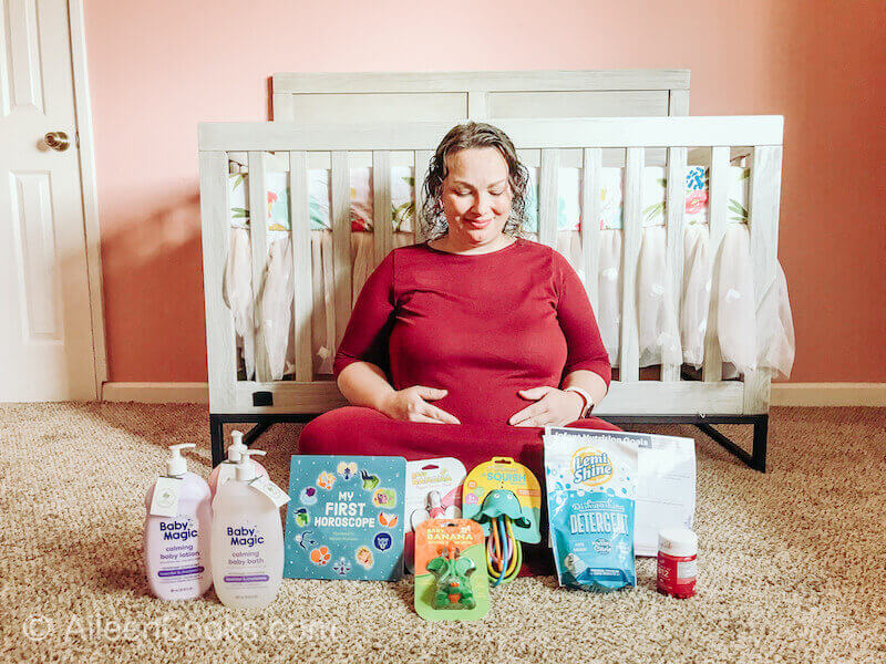 A pregnant woman in a red dress holding her belly in front of a crib and posing next to the contents of the New Mom and Baby Box.